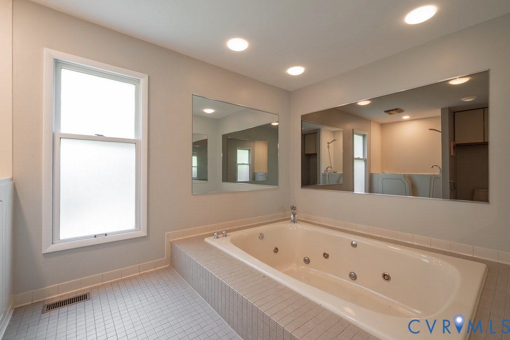 Jetted soaking tub with a tiled surround, recessed lighting, wall-mounted mirrors, and an opaque window