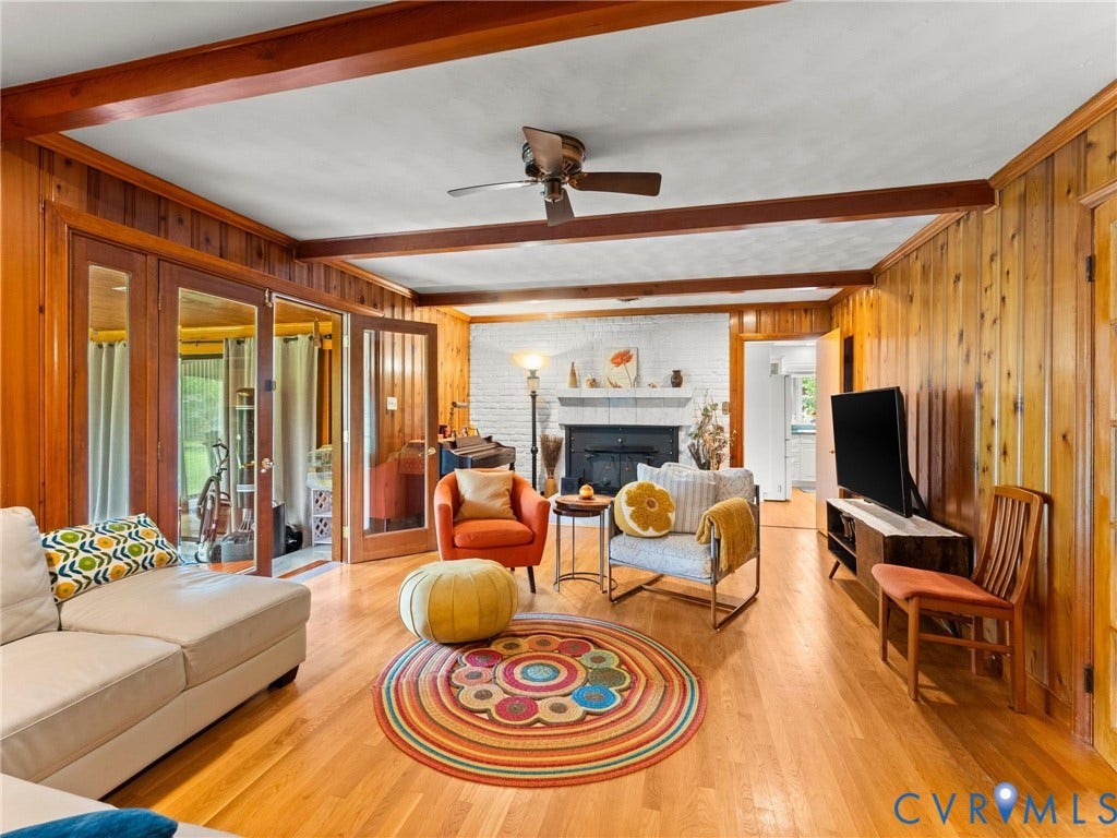 Living area featuring wood-finish flooring, wood wall paneling, exposed wood ceiling beams, and a brick fireplace with a white mantel