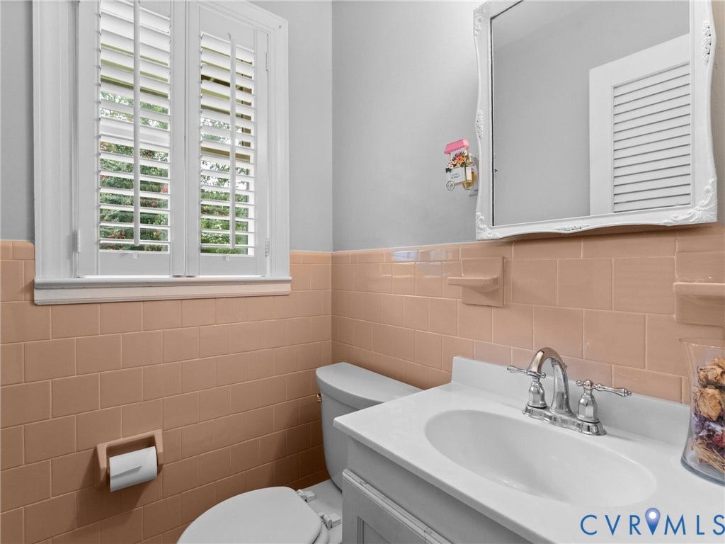 Bathroom featuring a window with white louvered shutters, a vanity with a white countertop and integrated sink, a chrome two-handle faucet, and pink subway tile wainscoting