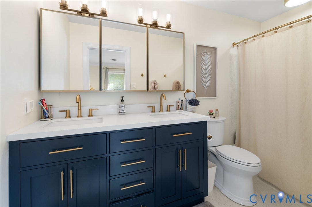 Dual vanity with a white countertop and navy cabinetry, brass-finish fixtures, triple mirrored medicine cabinet, and a white toilet