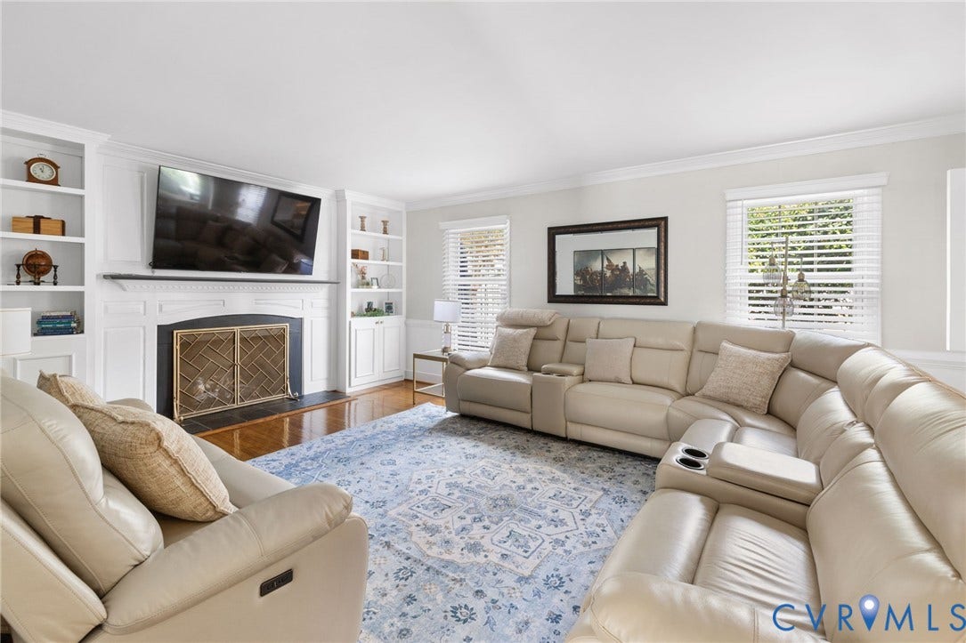 Bright and spacious living room featuring built-in cabinetry and shelving, a wall-mounted flat-screen television above a fireplace with decorative surround, hardwood flooring, large windows with plantation shutters, and a three-piece sectional seating arrangement with an ottoman