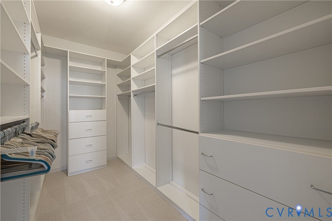 Custom Walk-in primary closet featuring white built-in shelving, multiple hanging rods, integrated drawers, and new carpeting