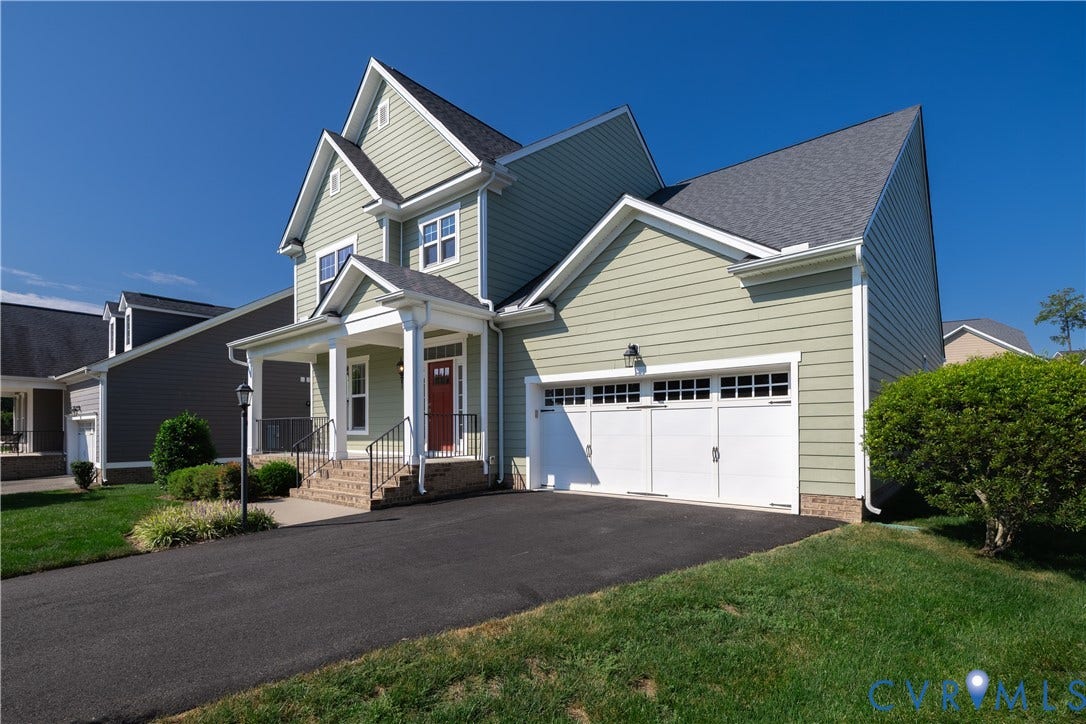 Light green siding exterior with a covered porch, brick stairs, and a two-car garage with a paved driveway