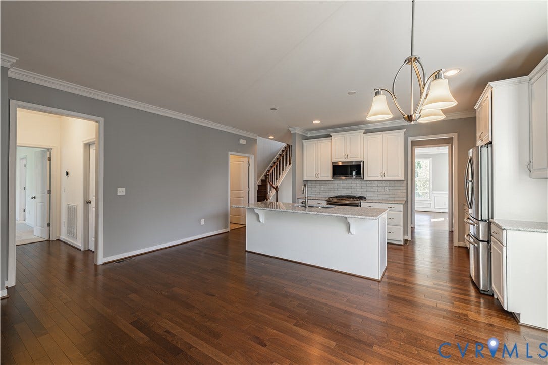 Hardwood flooring throughout, kitchen island with built-in sink, white cabinetry with crown molding, subway tile backsplash, and stainless steel appliances