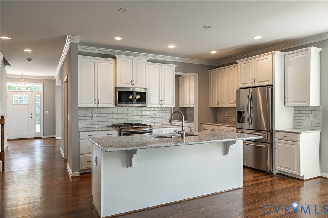 Wood flooring, white cabinetry, stainless steel appliances, subway tile backsplash, and a central island with a granite countertop
