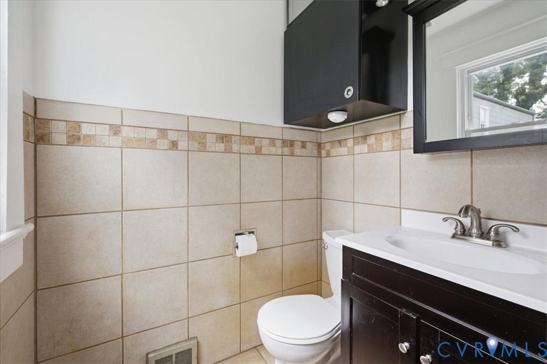 7232 Hopkins Road, North Chesterfield, VA 23237 Tile wainscoting with a decorative accent band, dark wood-finish vanity with an integrated sink, wall-mounted mirror, and an overhead storage cabinet