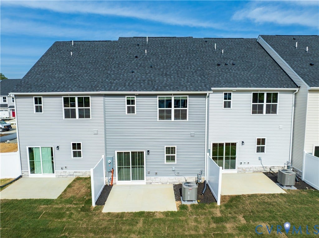 Rear exterior featuring light grey siding, a dark grey shingle roof, multiple windows, and sliding glass doors opening to concrete patios