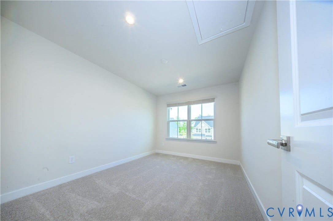 Recessed lighting fixtures, broad window with blinds, light-colored carpeting, white baseboards, and a white interior door with a brushed nickel lever