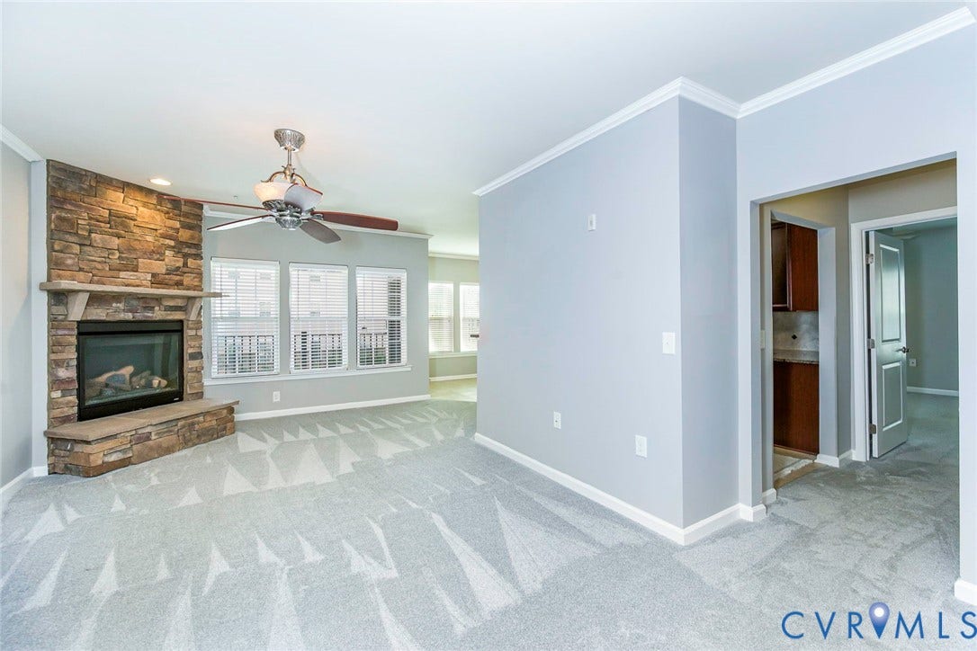Stone-clad fireplace with mantel, ceiling fan with integrated lighting, multiple windows, light-toned carpeting, and crown molding