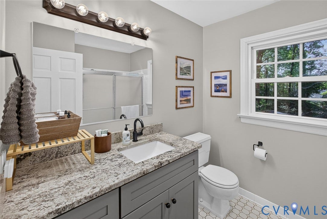 White hexagonal floor tiling, grey vanity with a built-in sink, stone-finish countertop, and a window with white trim