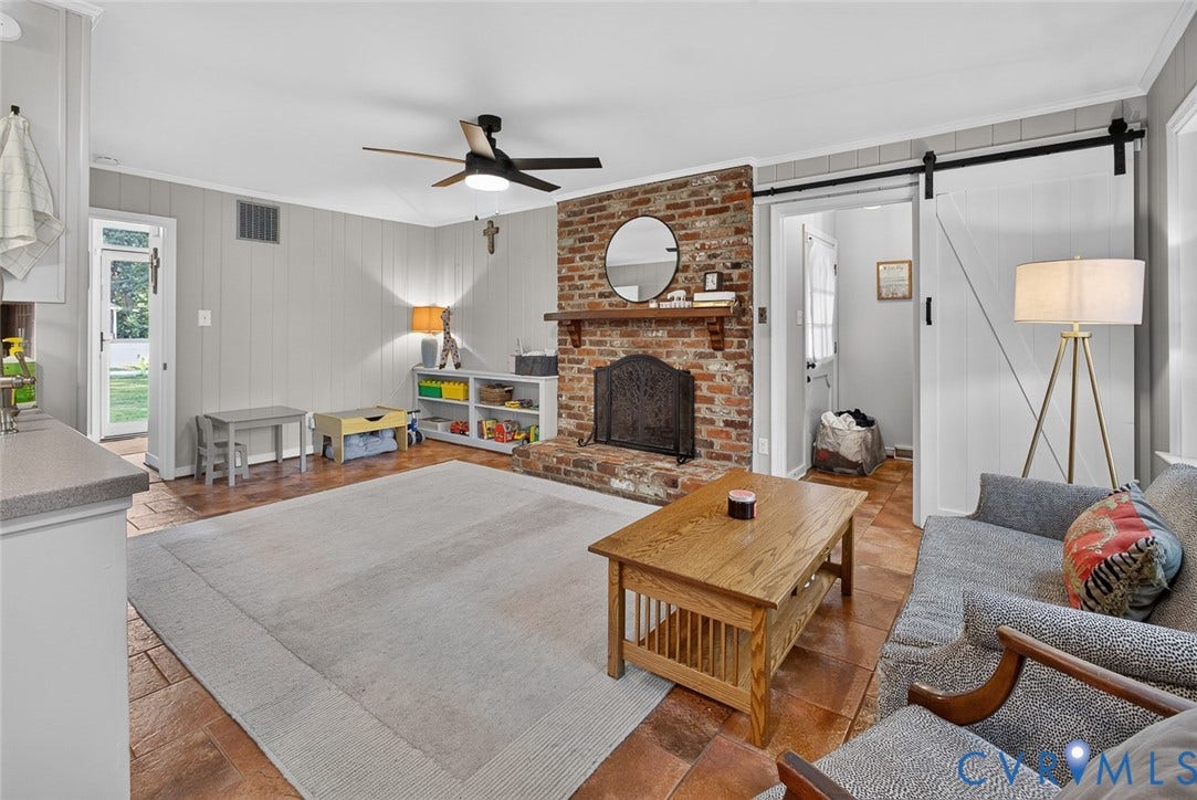 Exposed brick fireplace with a wood mantle, ceiling fan, wide plank wall paneling, tile flooring, and a white barn door