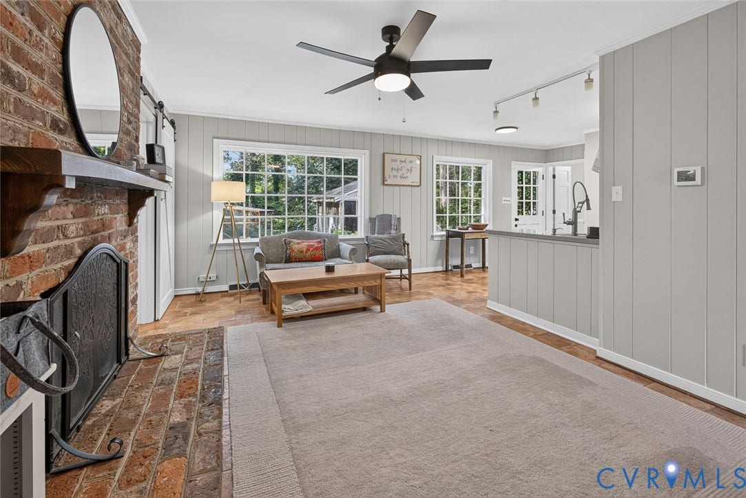 Brick fireplace with a wood-finish mantel, multiple large windows, wood-finish flooring, black ceiling fan, and white vertical paneled walls