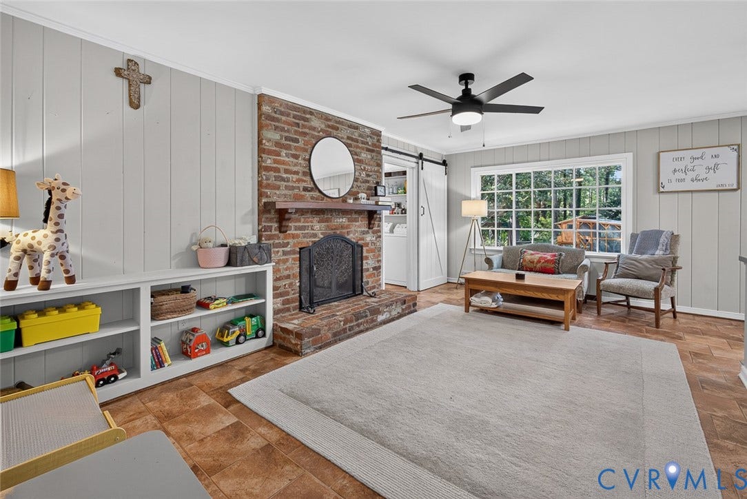 Brick fireplace with a wood mantel, paneled wall detailing, built-in shelving, a ceiling fan, and large multi-pane windows