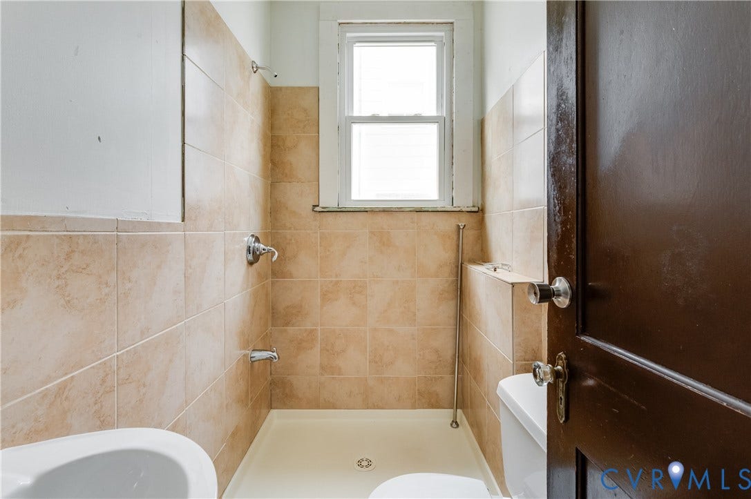 Tiled shower surround with chrome fixtures, window with privacy covering, and dark wood-paneled door