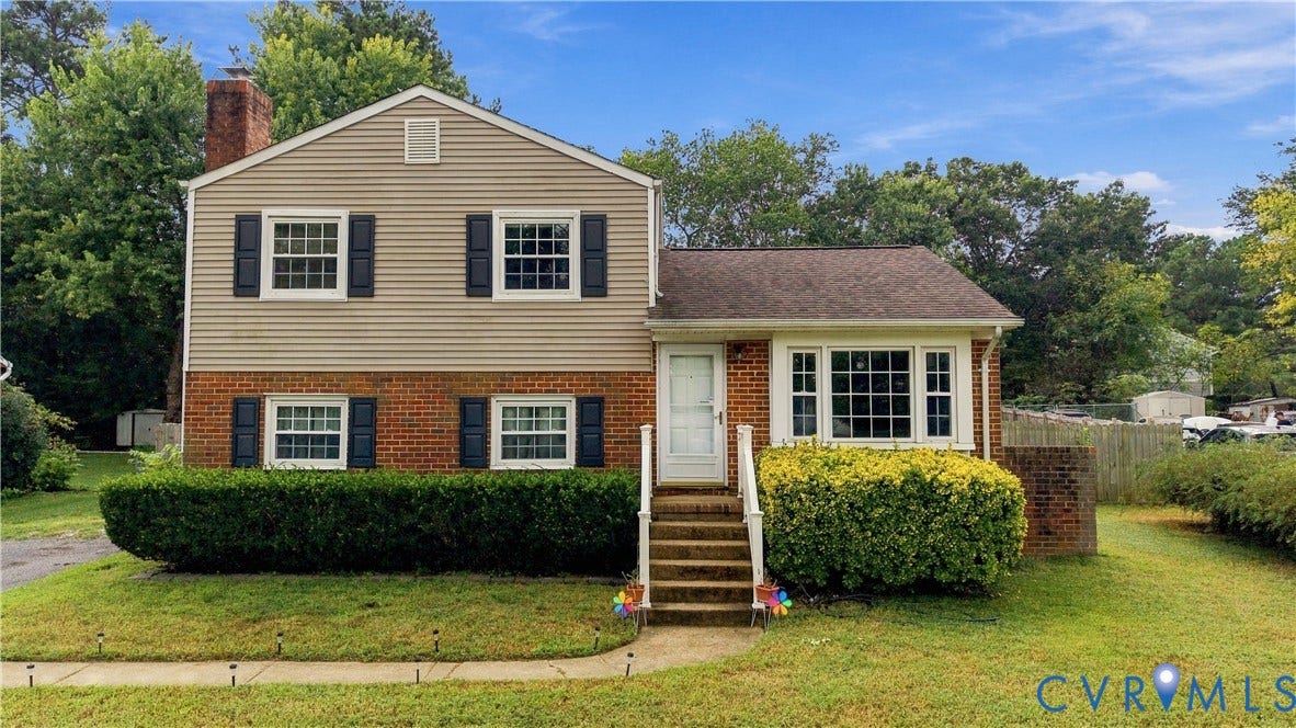 Brick and siding exterior with a prominent chimney, multiple windows featuring dark shutters, a front entry with steps and a handrail, and established front landscaping