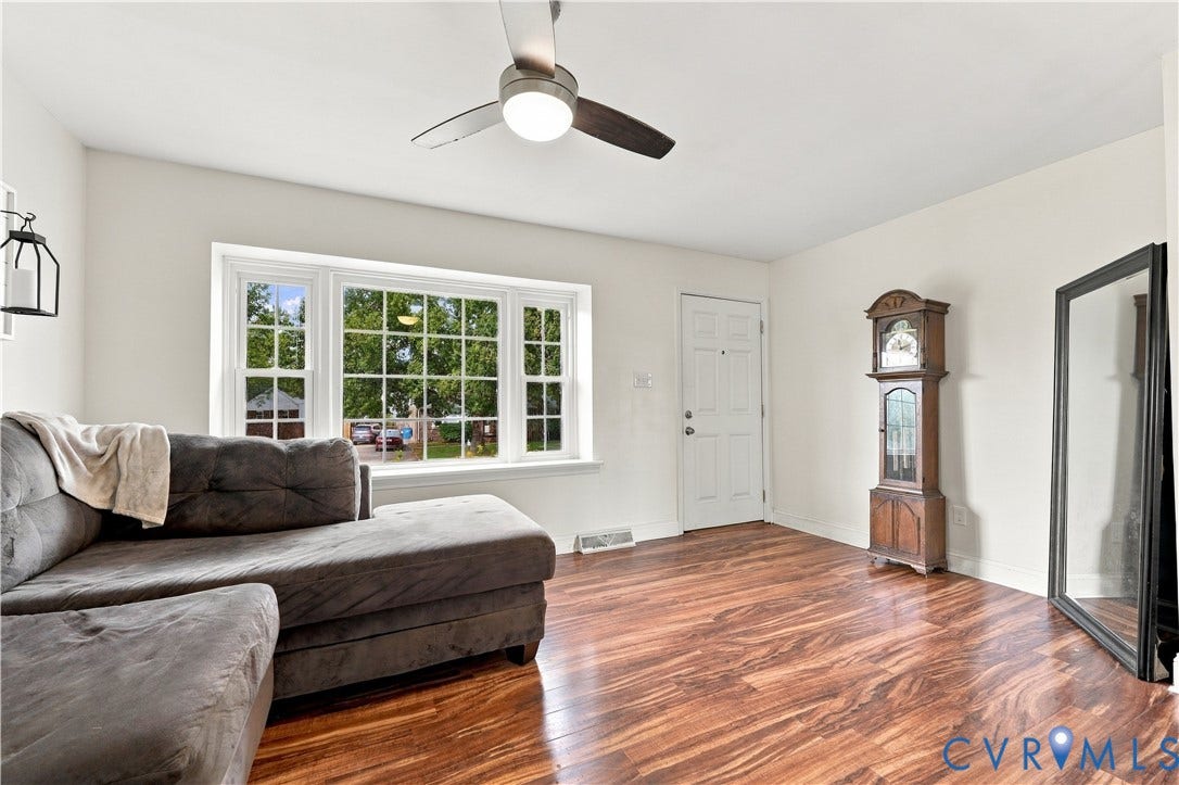 Spacious living area featuring wood-finish flooring, a ceiling fan with integrated lighting, and a bay window with multiple panes