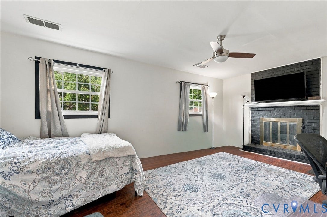 Wood-finish flooring, multiple windows, a ceiling fan, and a painted brick fireplace with a mantel