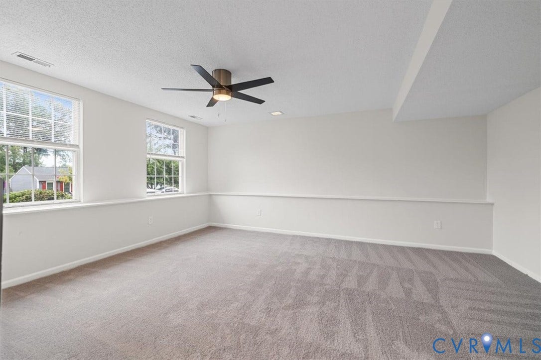 Bright, light-filled bedroom featuring multiple large windows with natural sightlines, neutral wall finishes with wainscoting detail, ceiling-mounted fan with integrated lighting, and plush carpeting throughout