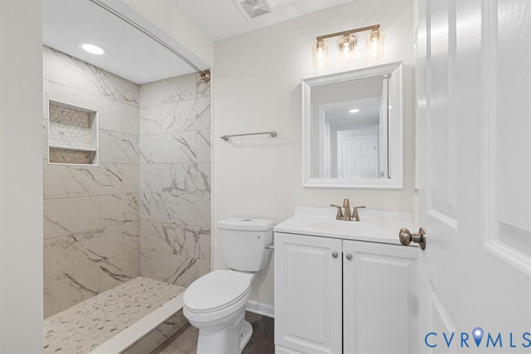 Walk-in shower with tiled walls, built-in shower niche, white vanity, and wall-mounted light fixture new shower curtain holder