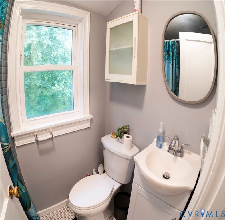 6706 Jahnke Road, Richmond, VA 23225 Single-hung window with white trim, wall-mounted storage cabinet with frosted glass panels, oval mirror with a gray frame, white pedestal sink with chrome faucet, and white hexagonal tile flooring