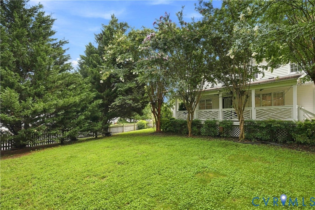 Expansive lawn area, mature evergreen trees, white picket fence, white exterior siding, and a covered porch with decorative railing