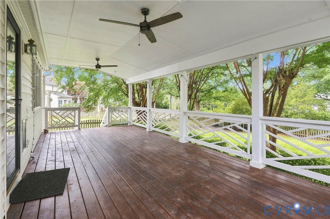 Expansive covered porch featuring a rich wood-finish deck, white railing with decorative cross-hatch design, and two ceiling fans