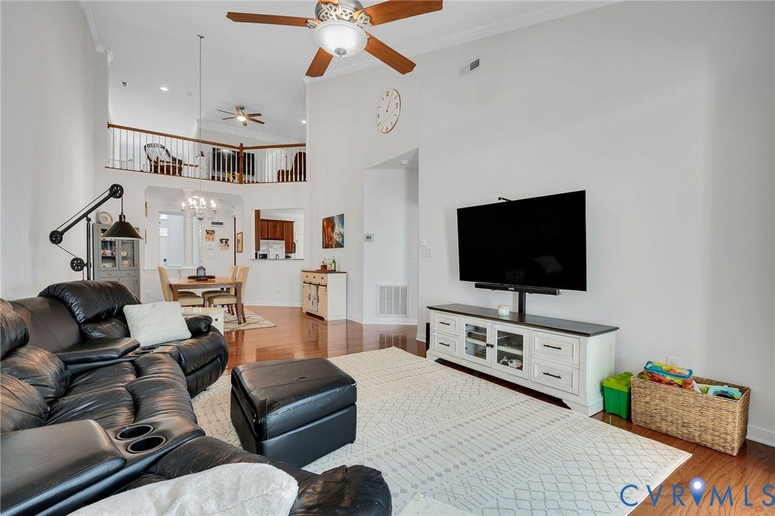 2811 Murano Way, The Villas At Hunton Park, Glen Allen, VA 23059 Open-concept living space featuring soaring ceilings with ceiling fans, a wall-mounted flat-screen television above a white media console with storage, wood-finish flooring, and a lofted mezzanine with built-in shelving visible above
