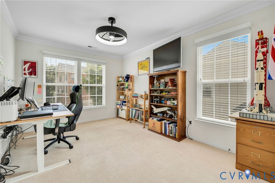 2811 Murano Way, The Villas At Hunton Park, Glen Allen, VA 23059 Bright, light-filled home office featuring multiple windows with plantation shutters, neutral carpeting, a modern ceiling fan with integrated lighting, and ample built-in wooden shelving and cabinetry for storage and display