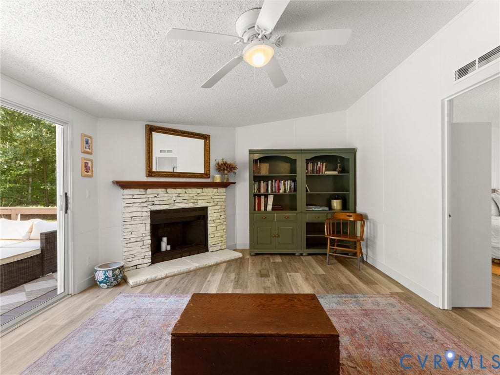 4554 Powhatan Lakes Road, Powhatan, VA 23139 Spacious living area featuring a wood-burning fireplace with stone surround and mantel, wide-plank wood-finish flooring, built-in shelving cabinetry with olive-toned painted finish, ceiling fan with integrated lighting, and glass sliding doors opening to outdoor space