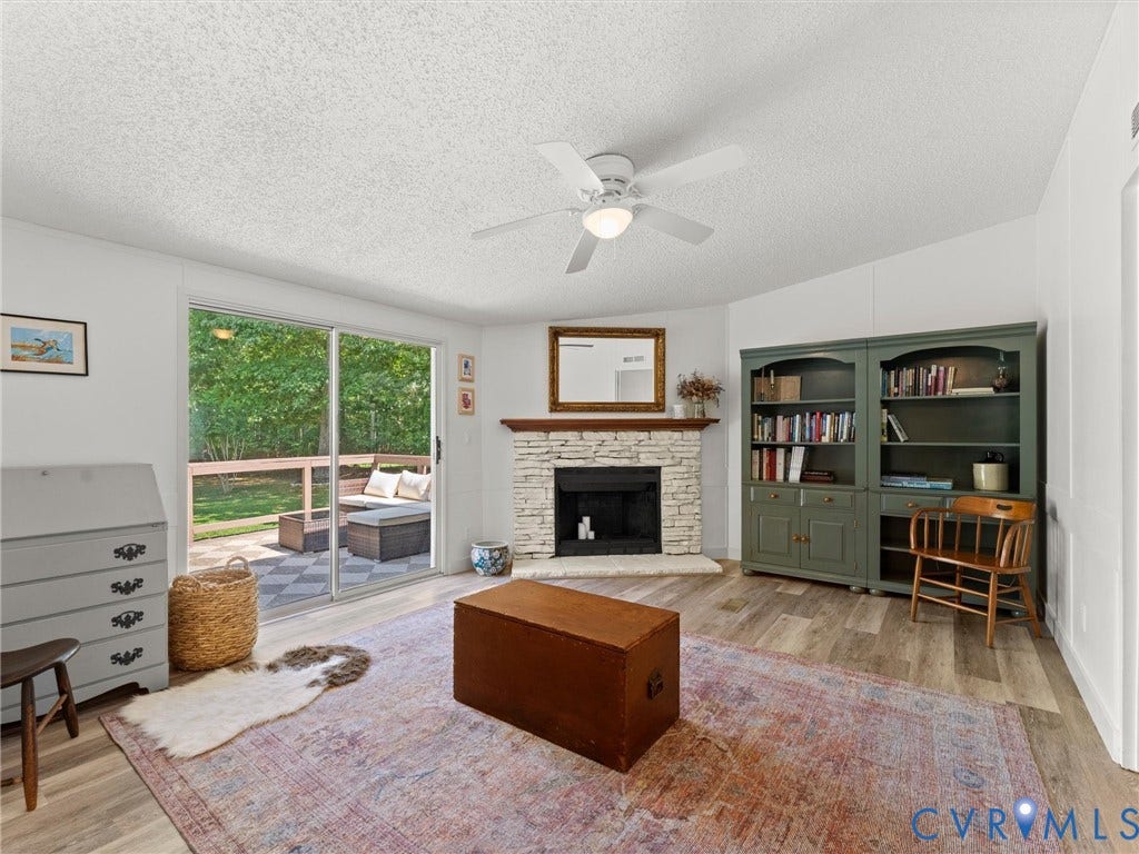 4554 Powhatan Lakes Road, Powhatan, VA 23139 Bright, open living room showcasing wood-finish flooring, a brick fireplace with mantel and hearth, built-in shelving with cabinet storage, ceiling fan, and sliding glass doors opening to a backyard with mature landscaping