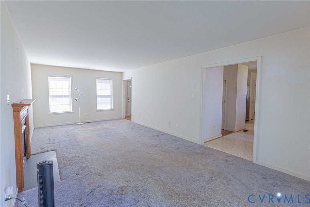 Spacious living area with a fireplace featuring a wood mantel, light-colored walls, and multiple windows