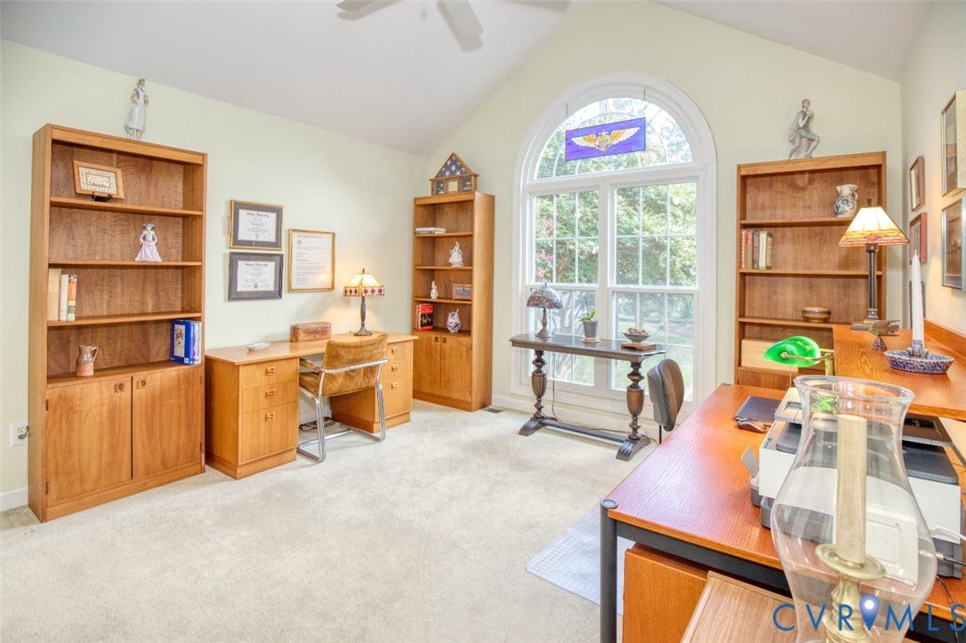 6311 Chiswick Park, Williamsburg, VA 23188 Second 1st floor bedroom or office suite featuring multiple wood cabinetry units with shelving and storage, abundant natural light from an arched transom window, wood-finish desk surfaces, ceiling fan, and neutral carpeting throughout the expansive room