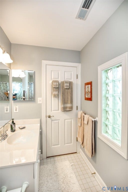 6311 Chiswick Park, Williamsburg, VA 23188 Bright, galley-style bathroom featuring a ceramic tile floor, white six-panel door with frosted glass inset, dual vanity with integrated sink and faucet, recessed ceiling lighting, and a window with decorative glass panes providing natural light