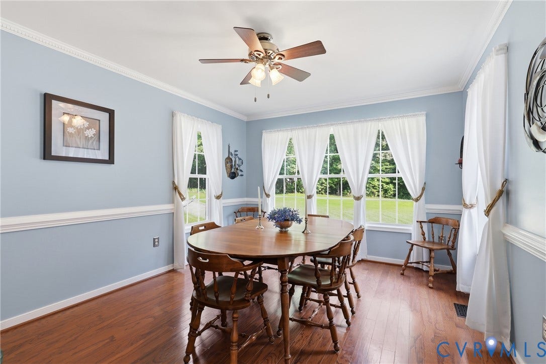 5295 Chimney Springs Drive, Saunders Acres, Goochland, VA 23063 Elegant dining room bathed in natural light from a series of multi-pane windows, featuring hardwood flooring, soft blue walls with white wainscoting, a ceiling fan with brushed metal finish, and crown molding throughout
