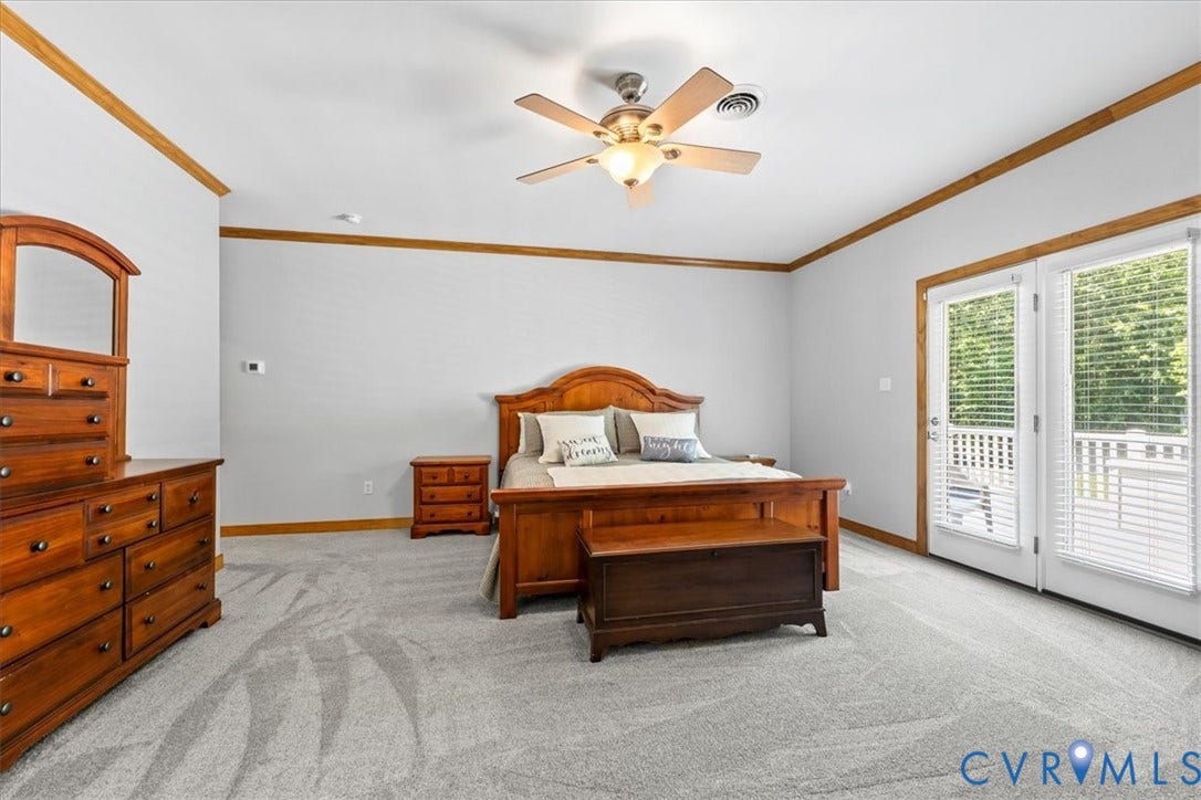 3060 Moyer Road, Powhatan, VA 23139 Spacious primary bedroom featuring wood-finish trim throughout, hardwood flooring, ceiling fan with integrated lighting, dual nightstands with wood construction, and glass sliding doors opening to a covered balcony with railing