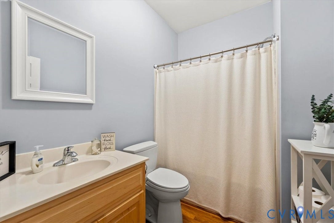 3060 Moyer Road, Powhatan, VA 23139 Bright bathroom featuring a wood-finish vanity with white countertop and integrated sink, white porcelain toilet, and a neutral fabric shower curtain