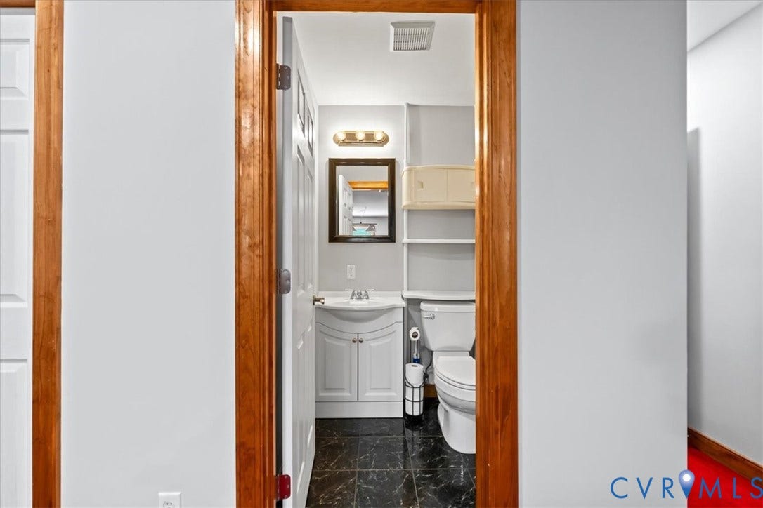 3060 Moyer Road, Powhatan, VA 23139 Wood-trimmed doorway leading to a powder room with black tile flooring, a white vanity with an integrated sink, a wall-mounted mirror, and a toilet