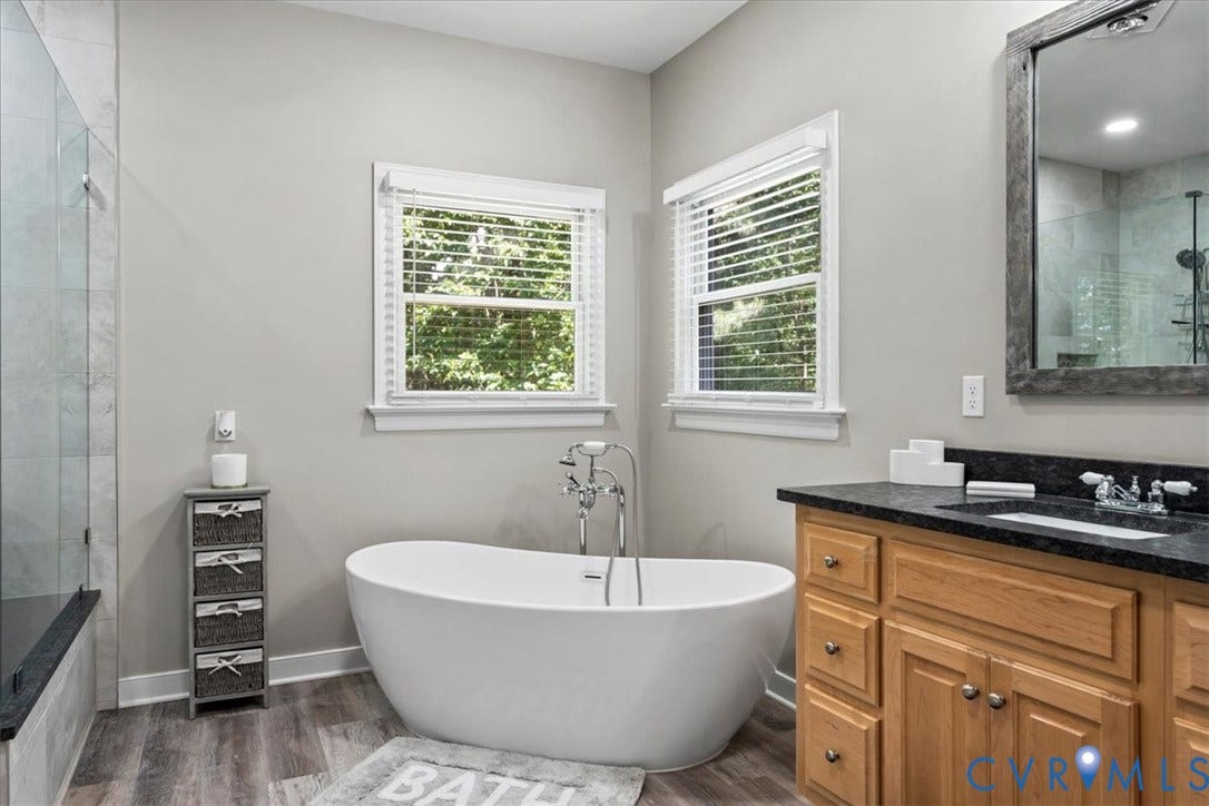 3060 Moyer Road, Powhatan, VA 23139 Spa-inspired primary bathroom featuring a freestanding soaking tub, dual vanity with wood cabinetry and dark countertops, frameless glass shower enclosure, and abundant natural light from two casement windows