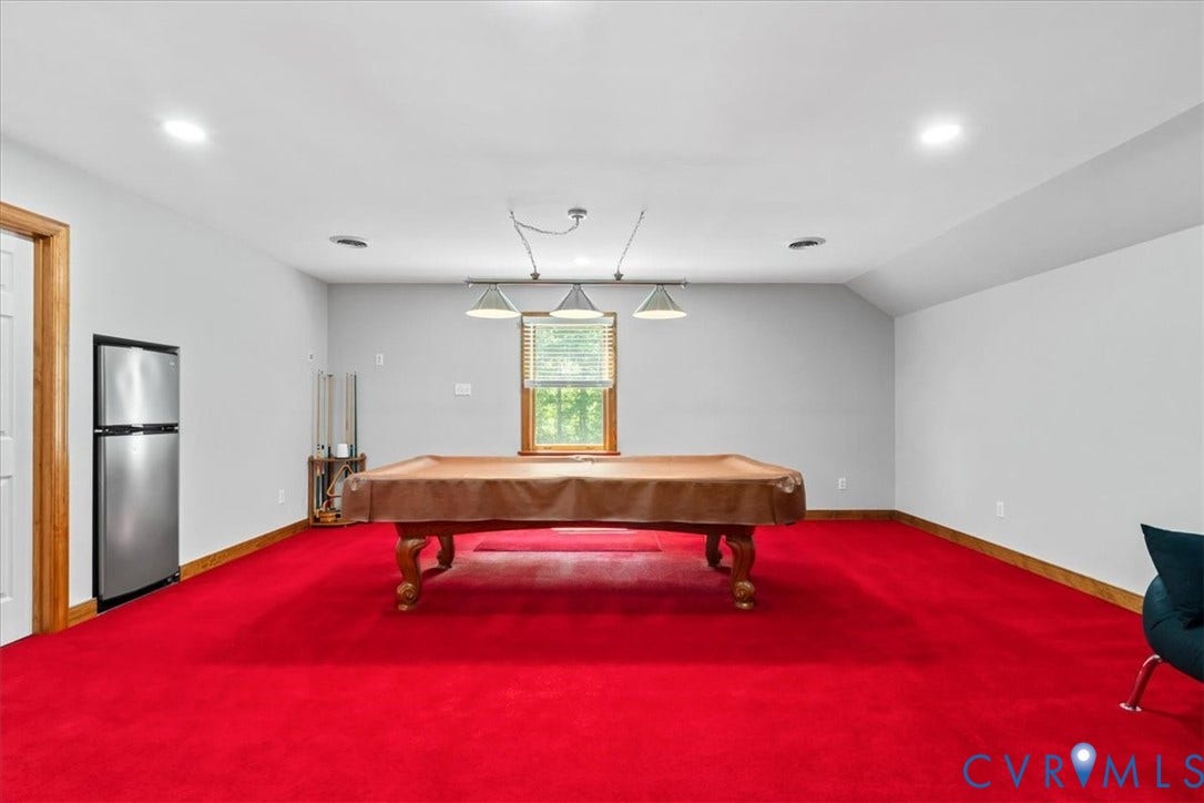 3060 Moyer Road, Powhatan, VA 23139 Spacious recreational room featuring vibrant red broadloom carpeting, wood-finish pool table with carved legs, recessed ceiling lighting, natural light from a large window, and convenient access to a refrigerator and adjacent utility spaces