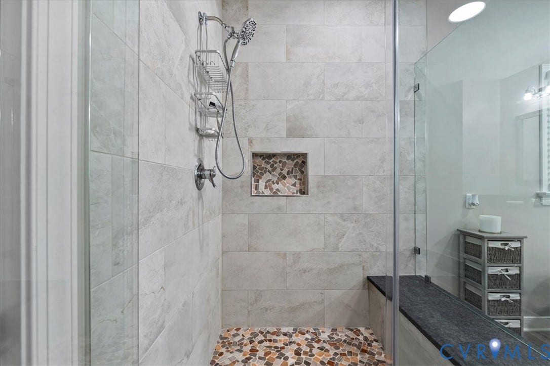 3060 Moyer Road, Powhatan, VA 23139 Spa-inspired walk-in shower featuring large-format marble-look tile walls, a rainfall showerhead with handheld spray fixture, recessed shelving niche, and a pebble stone accent floor