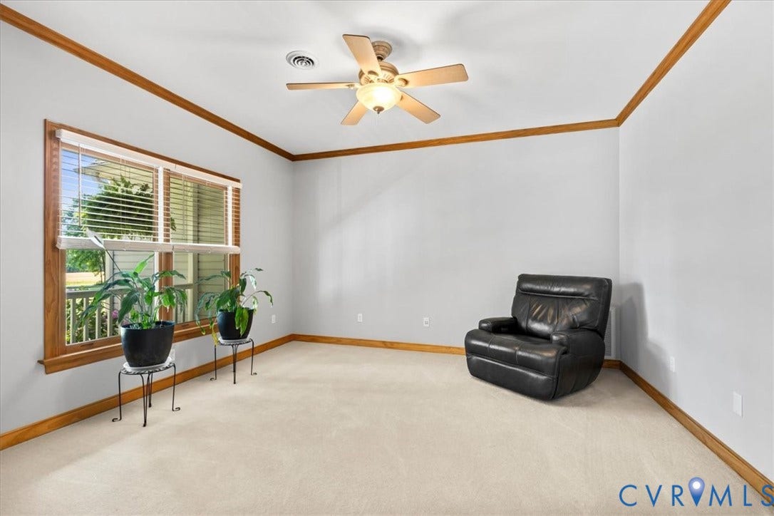 3060 Moyer Road, Powhatan, VA 23139 Bright corner room featuring wood-trimmed crown molding, ceiling fan with integrated lighting, expansive windows with wood-frame casements and plantation shutters, and neutral carpet flooring