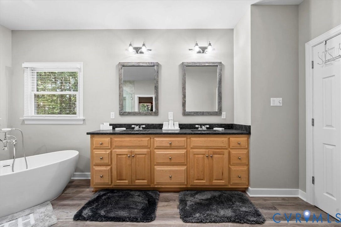 3060 Moyer Road, Powhatan, VA 23139 Spa-inspired primary bathroom featuring a freestanding soaking tub, dual-sink vanity with warm wood cabinetry and black countertops, paired sconce lighting above each mirror, large windows with natural light, and wood-finish flooring throughout