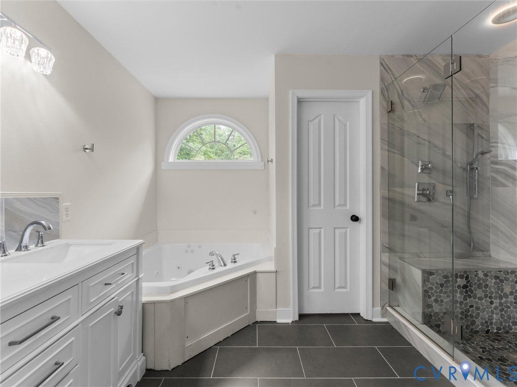 5017 Sadler Glen Place, Glen Allen, VA 23060 White vanity with dual sinks, corner jetted tub, arched window, walk-in shower with glass enclosure, and dark tile flooring