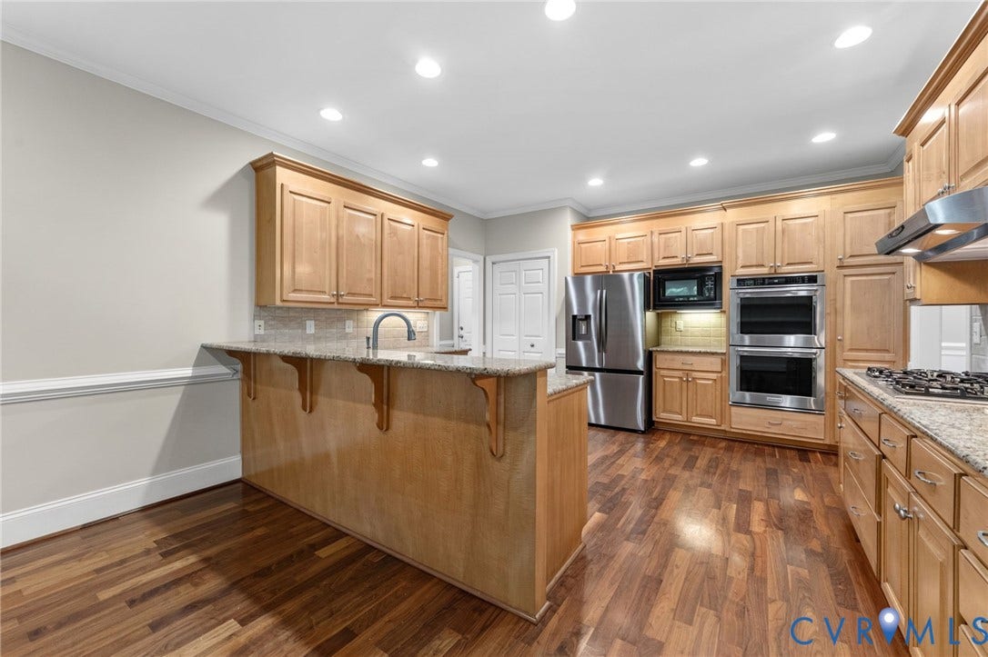 Spacious kitchen featuring warm wood cabinetry, stainless steel appliances including a gas cooktop and double wall ovens, a large center island with wood-finish base, wood-finish flooring, recessed ceiling lighting, and granite countertops with an undermount sink