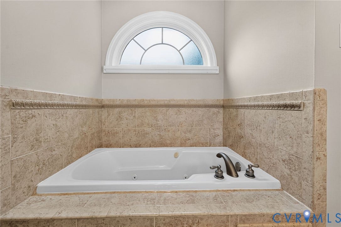 Built-in soaking tub with bronze-finish fixtures, surrounded by tiled decking and wall accents, under an arched transom window