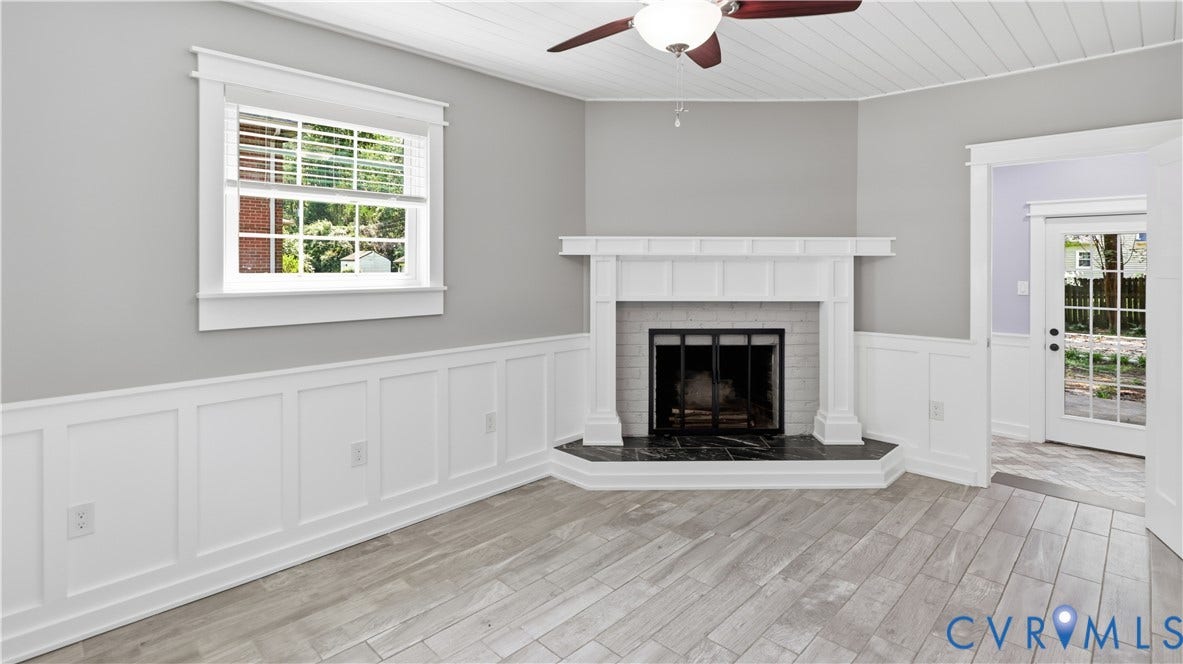 Fireplace with stone hearth, wainscoting, wood-finish flooring, ceiling fan, and white trim