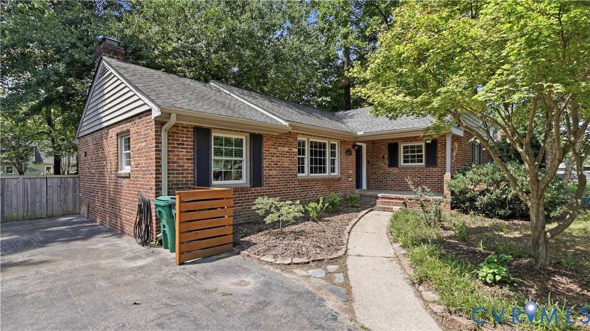 Brick exterior with dark shutters, covered front porch, concrete walkway, and mature trees
