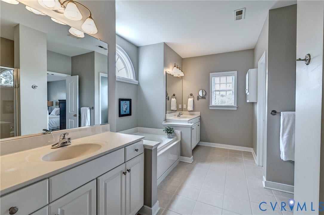 Dual vanity with white cabinetry, built-in soaking tub, arched window, and tile flooring