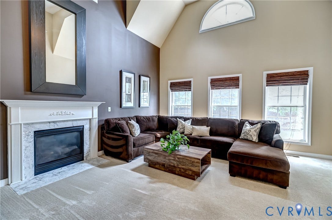 Soaring vaulted ceilings with an arched window, a fireplace with a white mantel and marble surround, and multiple windows with blinds