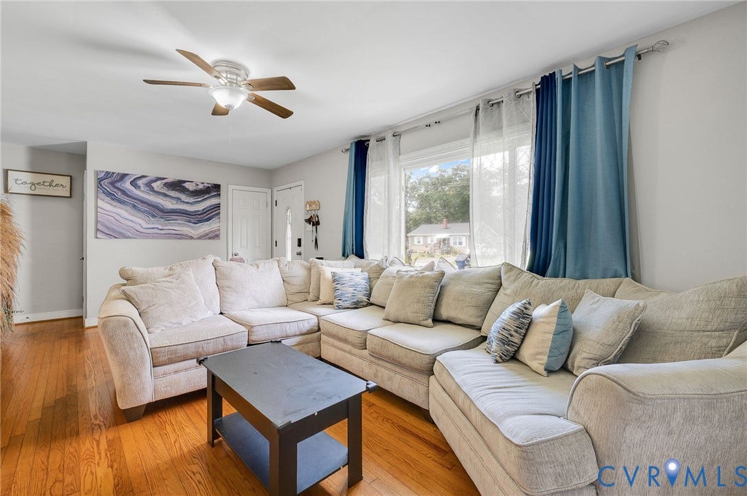 4819 Arundel Avenue, Chesterfield, VA 23234 Bright living room showcasing warm wood-finish flooring, expansive seating areas with neutral upholstery, large windows with blue drapery panels, ceiling fan with integrated lighting, and abundant natural light from multiple windows